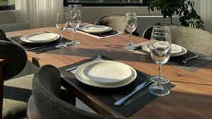 Mesa de comedor lista para cuatro personas, con copas de vino, platos y cubiertos, ubicada cerca de un ventanal con vistas a un bosque.