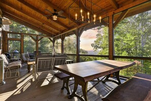 A covered outdoor area with a wooden table and benches, a ceiling fan, and a view of trees.