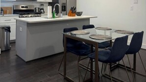 Een moderne keuken met een centraal eiland, een eetgedeelte met een glazen tafel en blauwe stoelen, en een roestvrijstalen fornuis.