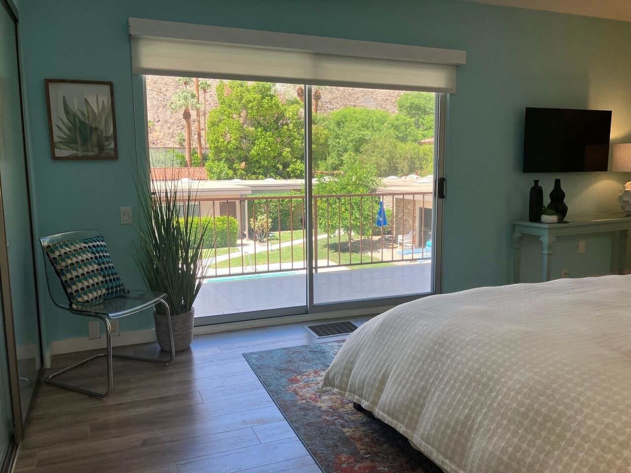 Une chambre avec un grand lit, une chaise, une plante en pot et une vue sur un balcon aménagé avec du mobilier d’extérieur.