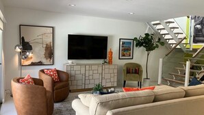 A modern living room with a flat-screen TV, two brown leather armchairs, a beige sofa, and a staircase with a glass railing. - 2-bedroom condo in Historic Tennis Club (Palm Springs)