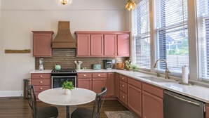 A modern kitchen with pink cabinets, a central island, and a large window with blinds. - The Estuary Indy - Your Private Sanctuary! (Indianapolis)