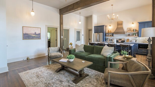 A spacious living room with a large green sofa, a wooden coffee table, and a patterned rug. A kitchen with dark cabinets and a stainless steel range is visible in the background. - The Estuary Indy - Your Private Sanctuary! (Indianapolis)