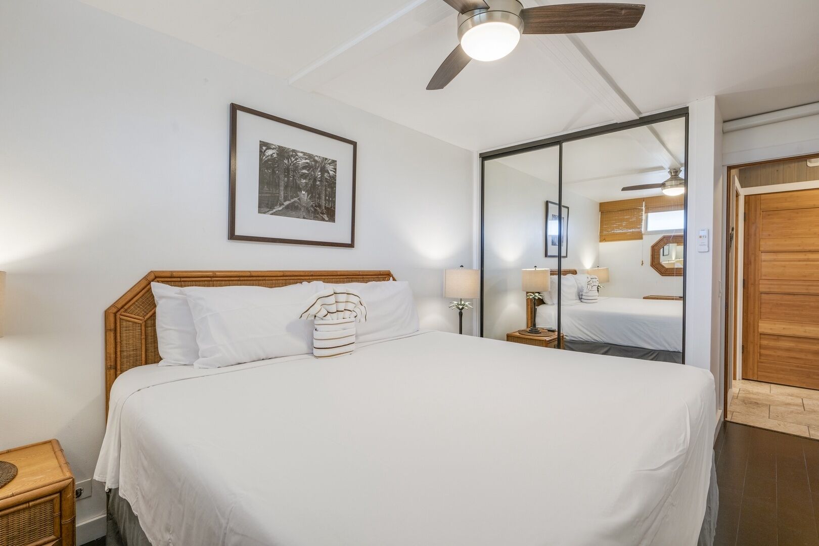 A bedroom with a large bed, a ceiling fan, a framed picture on the wall, and a wooden door.