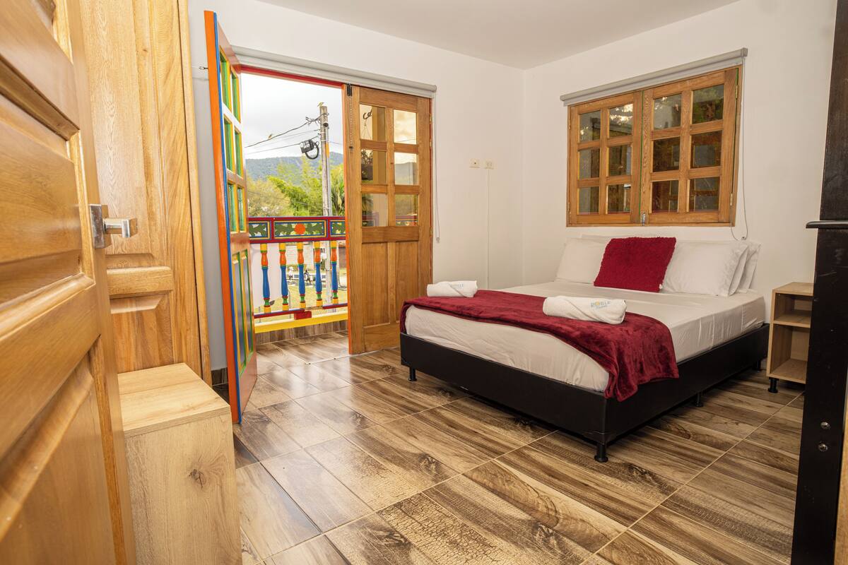 Una camera da letto con un letto in legno, una coperta rossa, un cuscino bianco, un comodino e una finestra con persiane di legno.