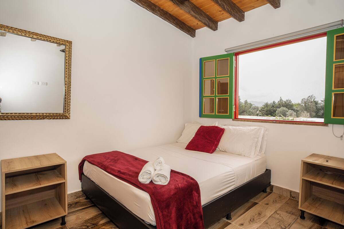 Una camera da letto con un letto, una coperta rossa, un paio di asciugamani arrotolati, un comodino in legno, uno specchio e una finestra con persiane verdi.