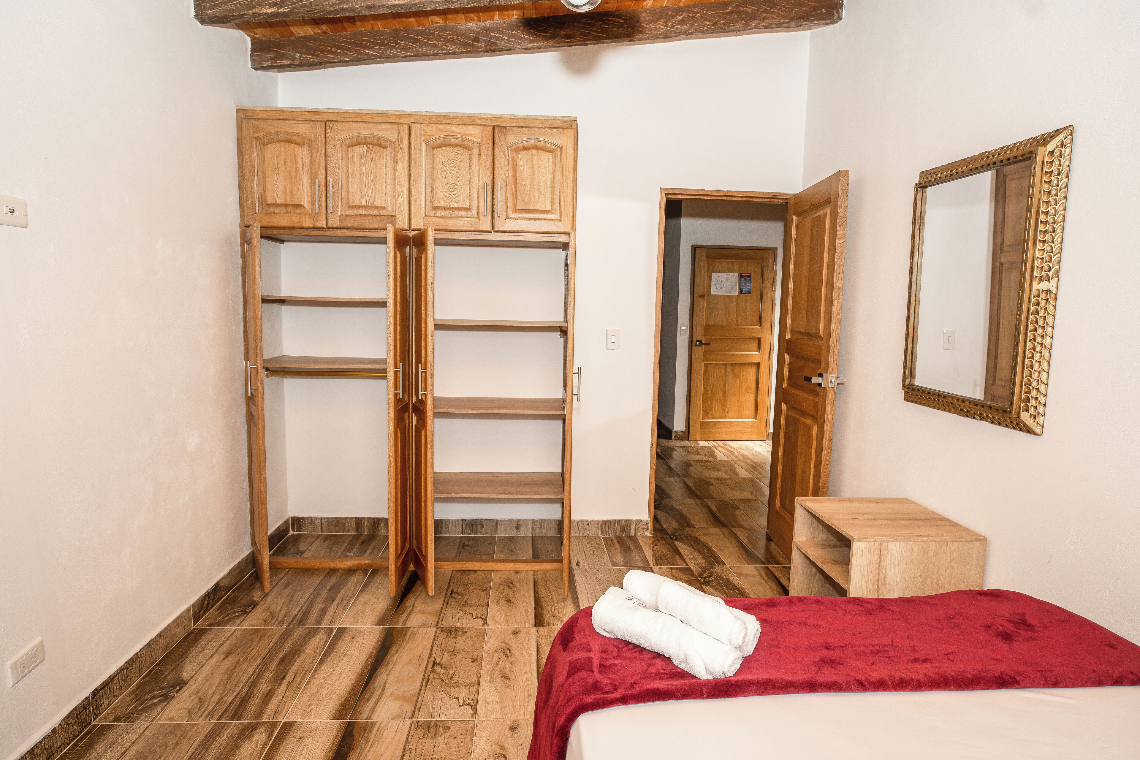 A bedroom with a bed, a red blanket, wooden wardrobe, and a mirror.