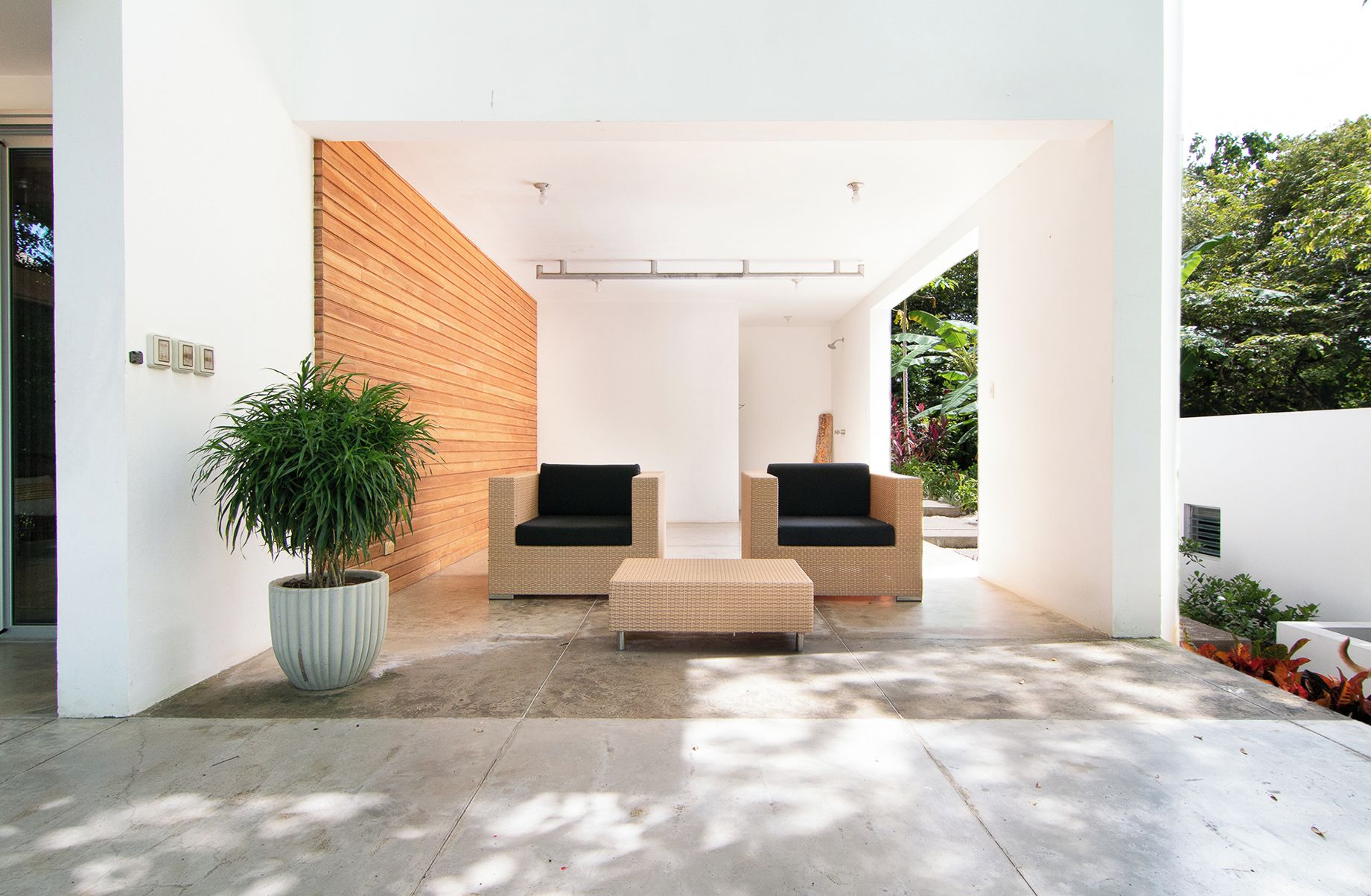 A modern outdoor seating area with two wicker chairs and a matching ottoman, a large potted plant, and a white wall with a wooden slat design.