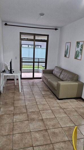 Una sala de estar con un sofá color beige, una mesa blanca y un amplio ventanal corredera que da a un balcón.