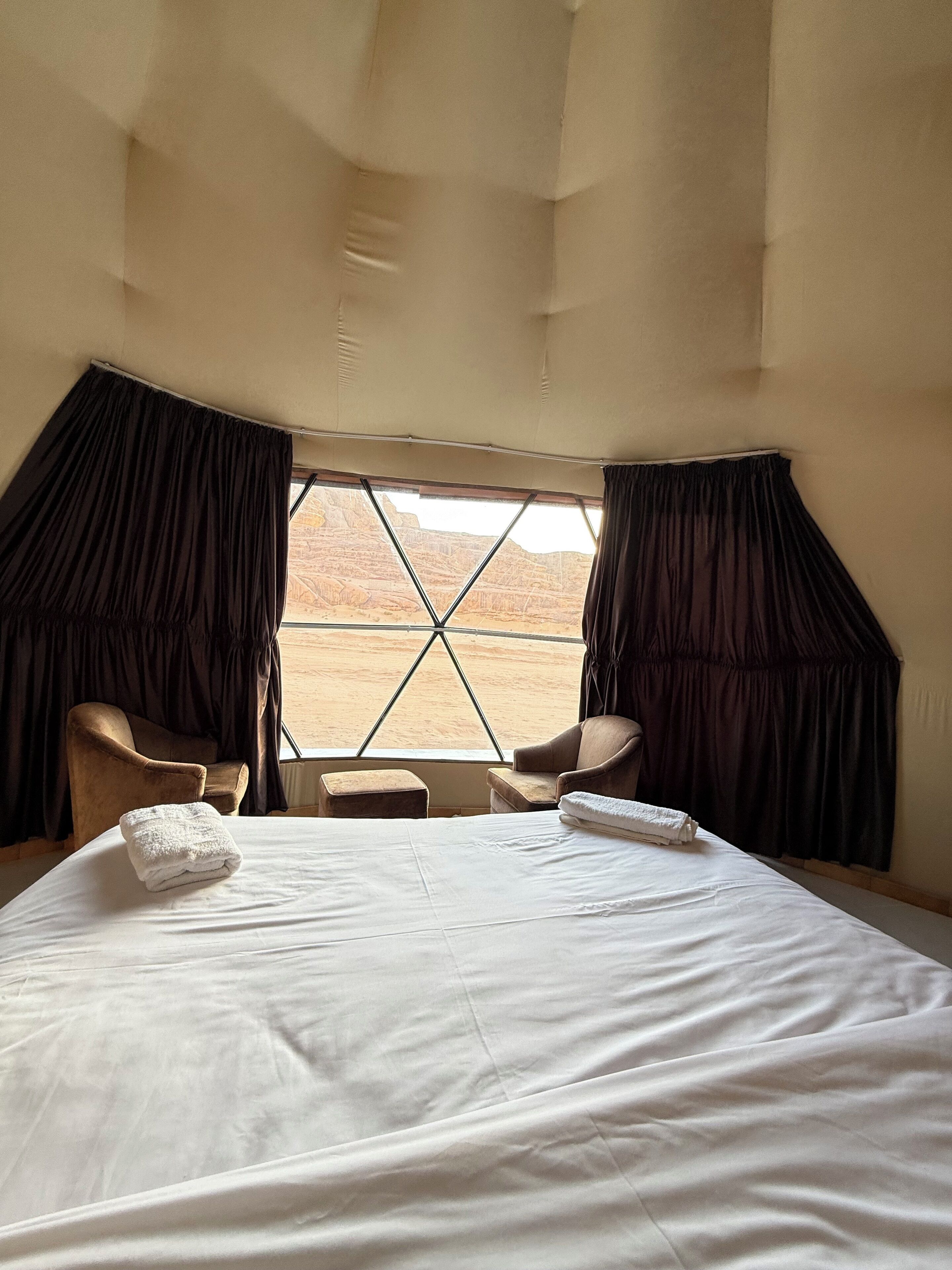 A hotel room with a large bed, two armchairs, a small table, and a view of a desert landscape through a window with curtains.