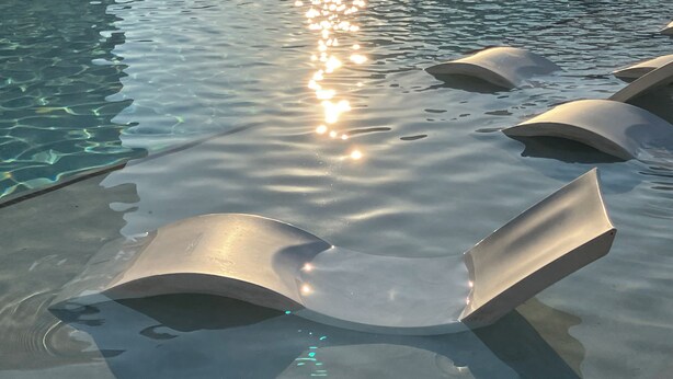 A pool with clear water and sun reflecting on the surface. There are three lounge chairs partially submerged in the water.