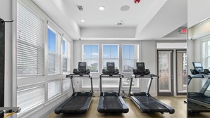 A gym with three treadmills aligned in a row, large windows with blinds, and a door with an exit sign.