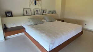 Een slaapkamer met een groot bed, een nachtkastje, een wandplank met ingelijste foto's en een airconditioner aan de muur.