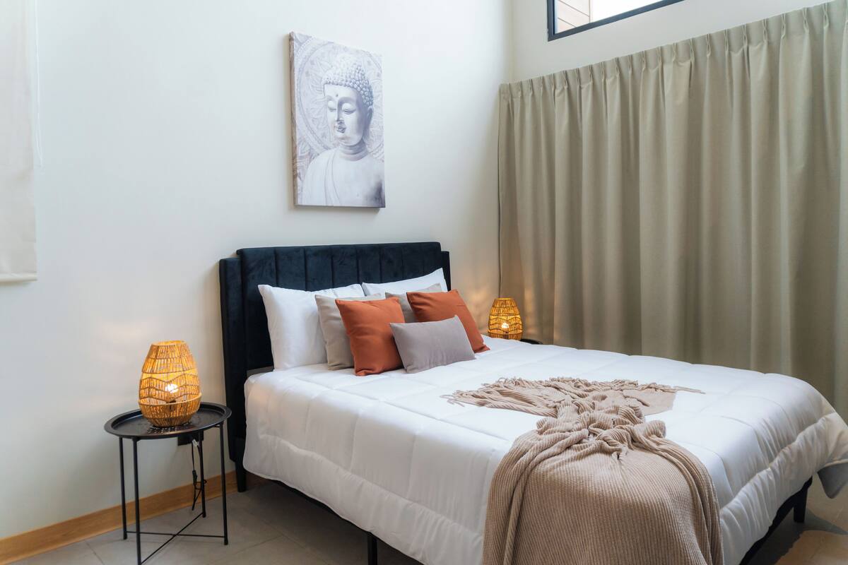 A neatly made bed with white bedding and a beige throw blanket, flanked by two bedside lamps on black side tables, a wall-mounted Buddha statue, and a large window with beige curtains.