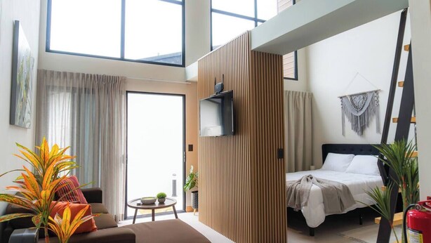 A modern hotel room with a large window, a bed with white linens, a wooden partition, and a mounted TV. - Sakura Haus (Turrialba)