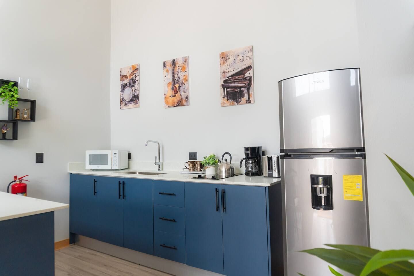 A modern kitchen with blue cabinets, a stainless steel refrigerator, a microwave, and a kettle. There are three framed artworks on the wall.