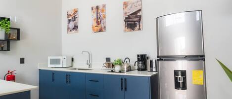 A modern kitchen with blue cabinets, a stainless steel refrigerator, a microwave, and a kettle. There are three framed artworks on the wall.