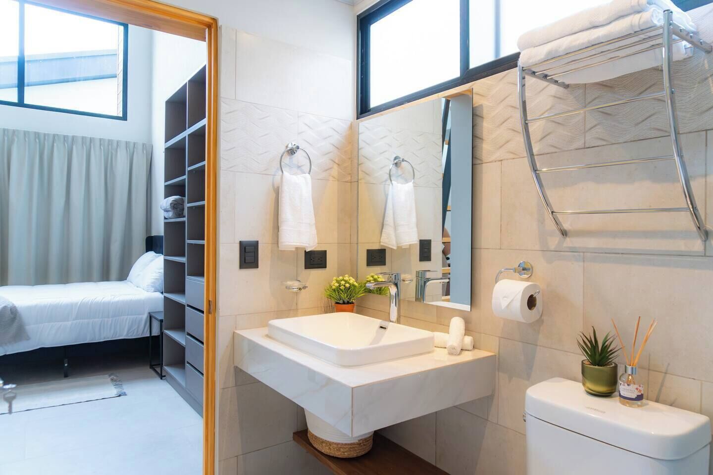 A modern bathroom with a large sink, mirror, and towel rack. There is a bed in the adjacent room.