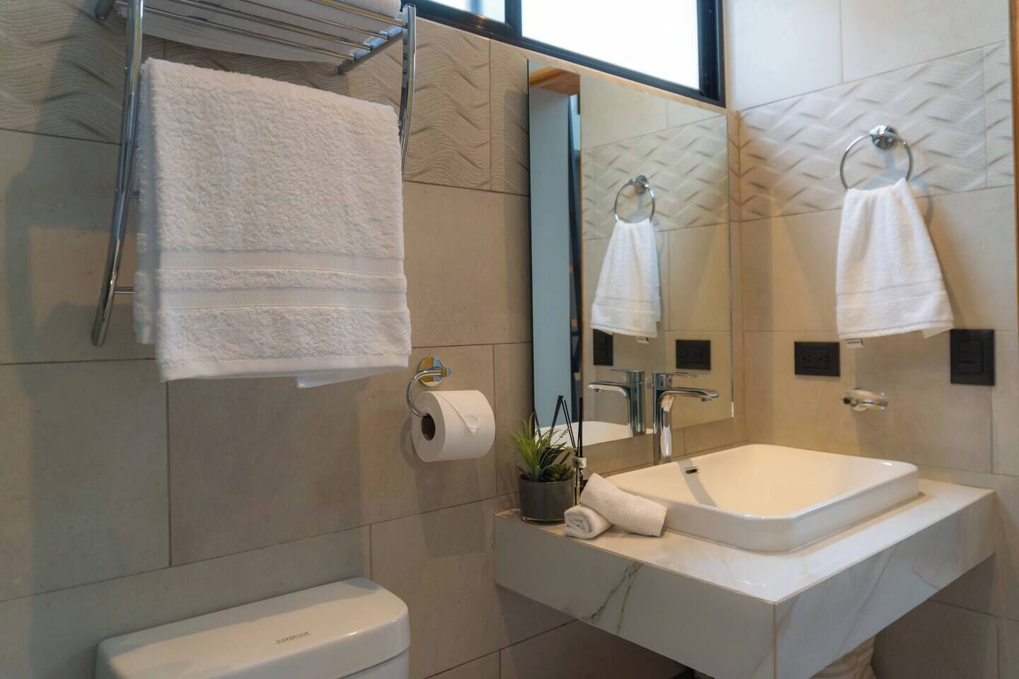 A modern bathroom with a white sink, a mirror, and a toilet. There are towels hanging on a rack and a roll of toilet paper on the wall.