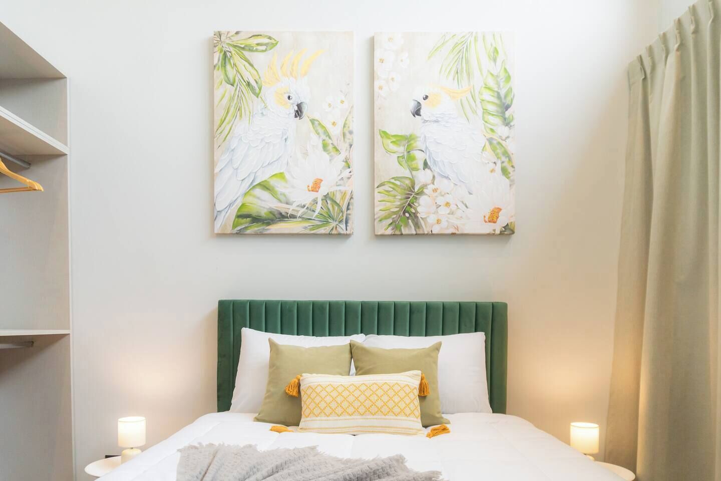 A bedroom with a green upholstered headboard, two white pillows, and a decorative pillow with a yellow pattern. There are two wall-mounted paintings of birds and foliage.