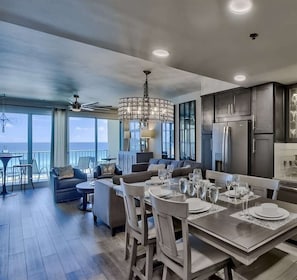 A spacious living area with a dining table set for six, a kitchen with dark cabinetry, and a balcony with a view of the sea.