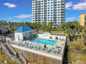 A pool area with a wooden deck, lounge chairs, and a building with a blue roof.