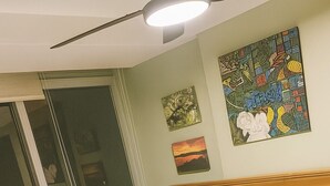 Un dormitorio con cama de madera, ropa de cama blanca, ventilador de techo y obras de arte en las paredes.