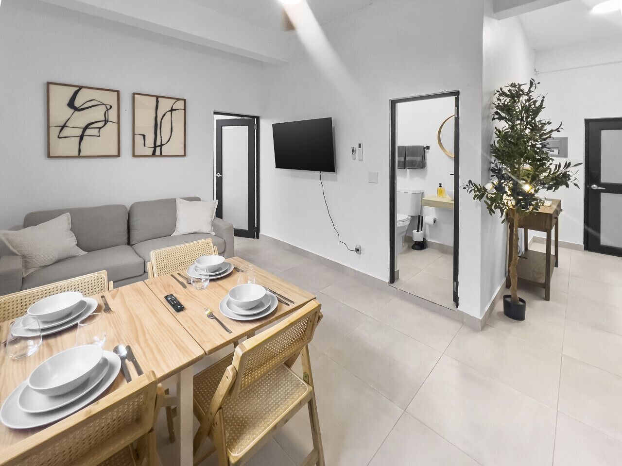 Ein moderner Essbereich mit einem Holztisch, gedeckt für vier Personen, einer grauen Couch und einem Fernseher an der Wand.