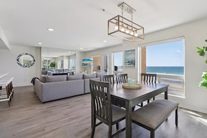 A modern living room with a gray sofa, wooden dining table, and chairs. There is a large window with a view of the ocean. - Modern Coastal Escape with Breathtaking Ocean View, Communal Pool and Barbecue (Imperial Beach)