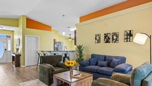 Una sala de estar con un sofá, sillones y una mesa de centro. Se ve una cocina al fondo, con un refrigerador y un lavamanos.