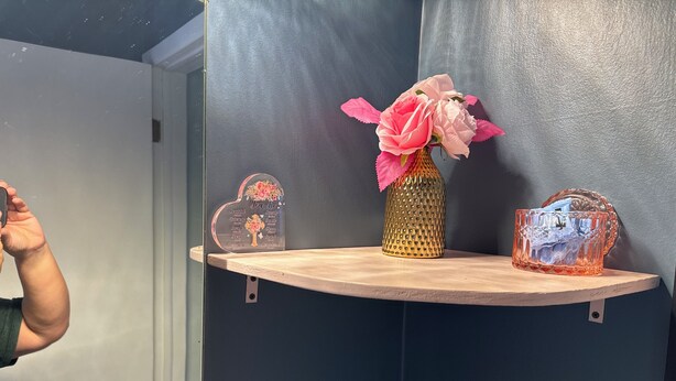 Un baño con un estante de madera que sostiene un jarrón con flores rosadas, un letrero decorativo y un recipiente de vidrio.