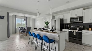 A modern kitchen with white cabinets, a stainless steel refrigerator, a microwave, and a stove. There are blue bar stools and a dining table with chairs. - Spacious 2BR Mickey Bedroom Lake View (Kissimmee)