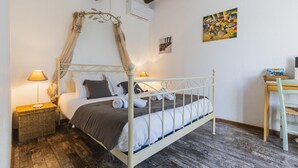 Een slaapkamer met een hemelbed, een nachtkastje met een lamp, een klein houten bijzettafeltje en een stoel.
