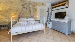 Une chambre avec un lit en métal blanc, une échelle en bois, un téléviseur posé sur une commode et un mur en pierre.