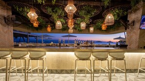Eine Bar mit Korbstühlen, hängenden Lampen aus Korbgeflecht und Blick auf den Sonnenuntergang über dem Meer.
