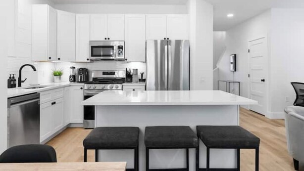 A modern kitchen with white cabinetry, stainless steel appliances, and a central island with bar stools. - Comfy Blue ~ King & Queen Beds w/Fast Wi-Fi (Austin)