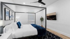 A modern bedroom with a four-poster bed, a ceiling fan, a mounted TV, and a potted plant. - Comfy Blue ~ King & Queen Beds w/Fast Wi-Fi (Austin)