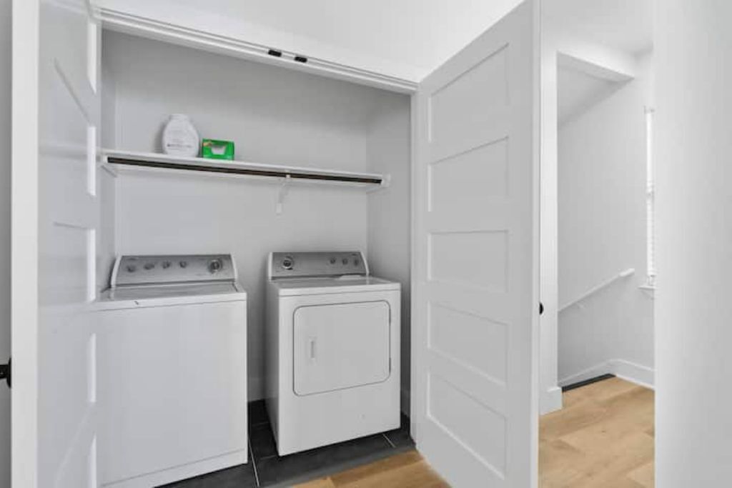 A laundry room with two white washers and dryers, a shelf with a green box, and a wooden floor.