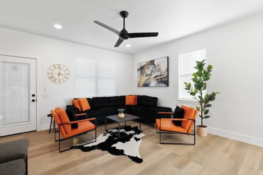 A modern living room with a black sectional sofa, orange armchairs, a cowhide rug, and a ceiling fan.