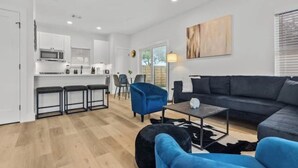 A modern living room with a dark grey sofa, a blue armchair, and a black ottoman. There is a kitchen area with white cabinets and a microwave. - ACL Ready ! ~ Near Downtown & Airport ~ Backyard! (Austin)