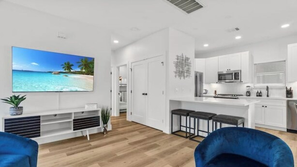A modern kitchen with white cabinets, a microwave, and a coffee machine. There is a bar area with stools and a large flat-screen TV displaying a beach scene. - ACL Ready ! ~ Near Downtown & Airport ~ Backyard! (Austin)