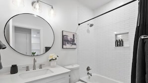 Een moderne badkamer met een wit wastafelmeubel, toilet en bad, een ronde spiegel en een douchegedeelte met een zwarte handdoek.