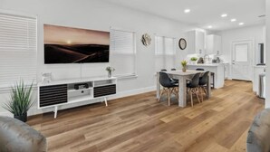 Un salon moderne doté d’un téléviseur à écran plat, d’un coin repas avec une table et des chaises, et d’une cuisine avec des meubles blancs et un plan de travail.