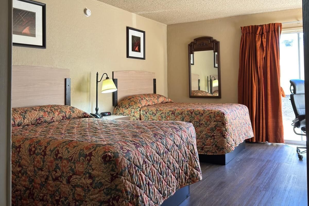 Habitación de hotel con dos camas, ventilador de techo, espejo y ventana con cortinas.