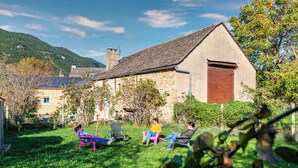 Un bâtiment rustique en pierre, doté d’une grande porte en bois, entouré d’un jardin luxuriant et surplombé d’un ciel dégagé.