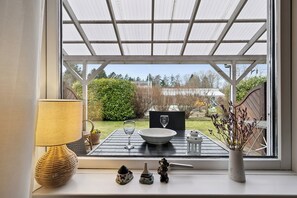 Ein Tisch mit einer Schale, einem Weinglas und einer Vase mit getrockneten Blumen, platziert auf einem Fensterbrett mit Blick ins Grüne.