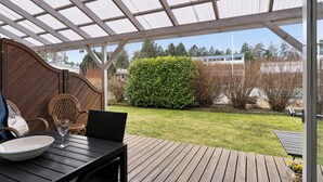 A covered outdoor patio with a table and chairs, a wooden deck, and a grassy yard with a hedge. - Holiday bliss by forest and Bornholm’s top beach (Nexø)