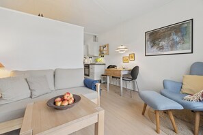Ein modernes Wohnzimmer mit einer Couch, einem Couchtisch mit Obstschale, einem Essbereich mit Schreibtisch und Stühlen sowie einer Küche im Hintergrund.