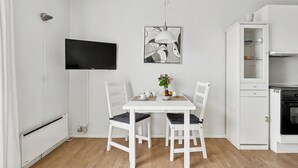 A dining area with a white table, two chairs, a flat-screen TV, a pendant light, and a cabinet with a microwave. - Pension Koch, lejlighed 14 (Gudhjem)