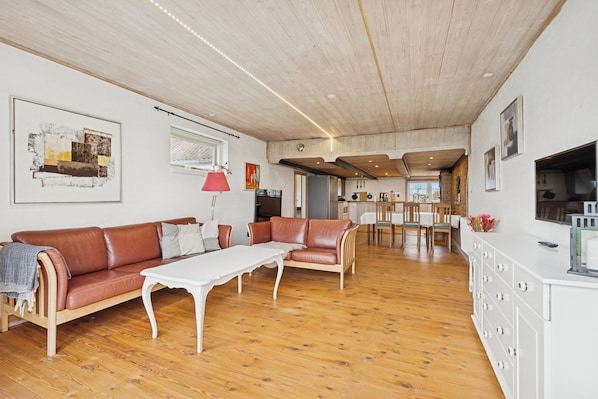 Un vaste espace de vie doté d’un plancher en bois, d’un canapé en cuir marron, d’une table blanche et d’un coin cuisine à l’arrière-plan.